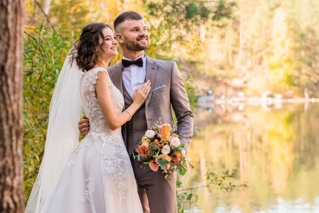 Stunning outdoor wedding couple smiling by lake with autumn trees.
