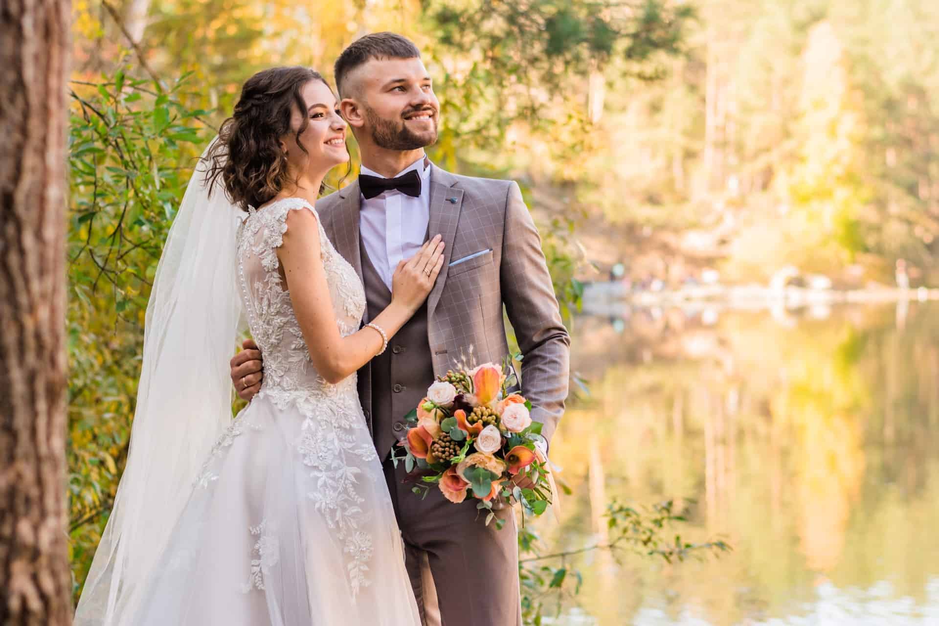 Stunning outdoor wedding couple smiling by lake with autumn trees.