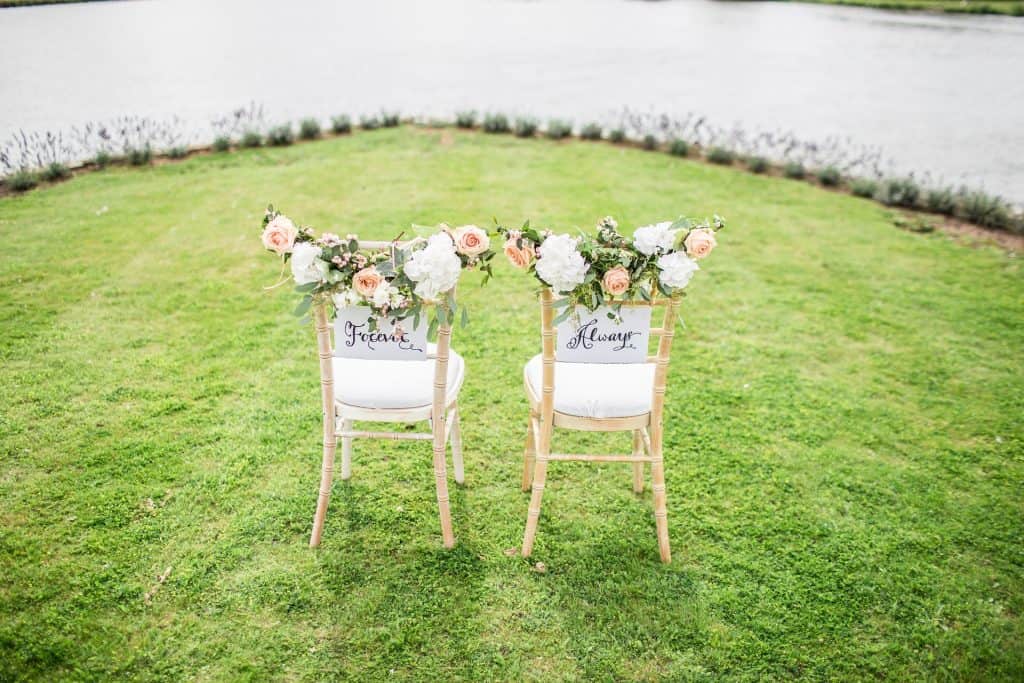 Elegant wedding aisle chairs decorated with floral arrangements on a lush green lawn with a waterfront view.