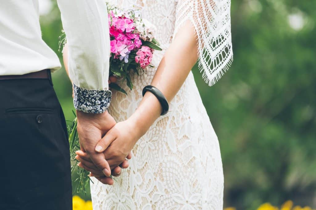Romantic couple holding hands with wedding dress and bouquet outdoors.