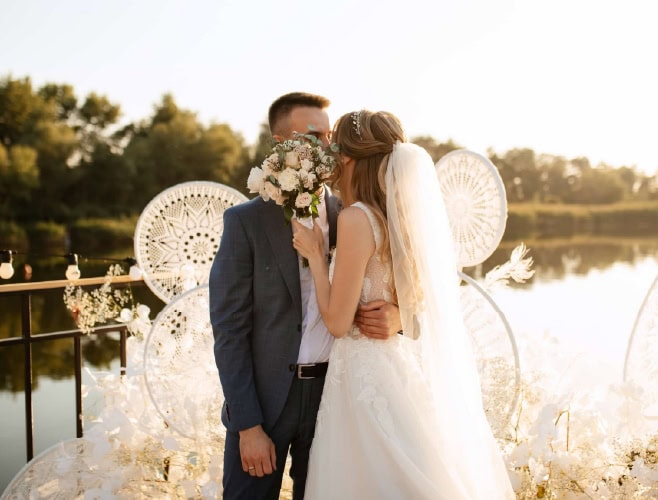 Elegant outdoor wedding ceremony with bride and groom by the lake, romantic sunset backdrop, with floral and dreamcatcher decor.